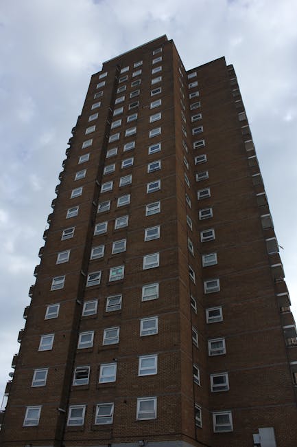 The image shows the exterior of a modern residential building with the name 'west/east' engraved on a concrete panel above an open parking area or entrance. The building features dark brickwork on eit