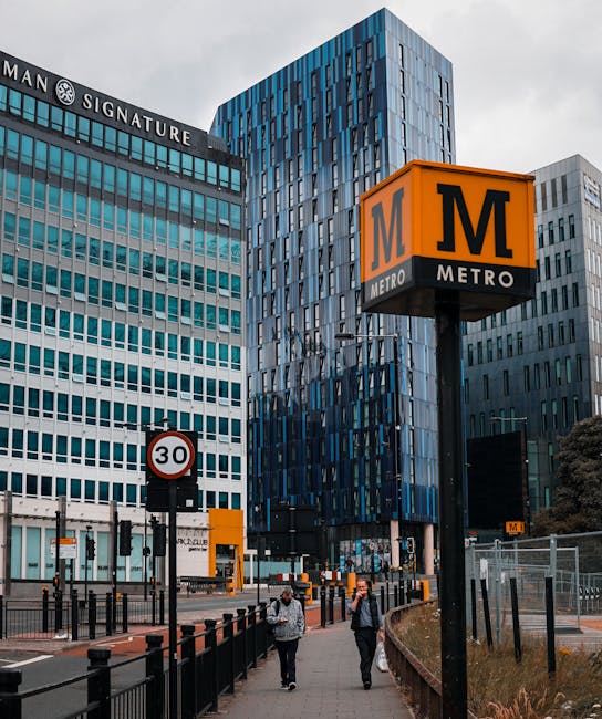 A modern urban scene featuring a London Underground metro sign with an orange background and black 'M' logo, positioned on a tall black pole near the sidewalk. Behind the sign are tall contemporary office buildings with glass facades, reflecting the sky and surrounding structures. One building displays a sign reading 'MAN SIGNATURE' at the top. The street scene includes a 30 mph speed limit sign, black metal railings along the pavement, and a few pedestrians walking past. The area appears to be a busy city centre location with a mix of commercial and transport infrastructure. This setting is relevant to house removals and moving services, illustrating access points and logistical considerations for furniture transport and home relocation near busy urban buildings, with the presence of loading zones and pedestrian pathways supporting efficient moving operations. The overall atmosphere is professional and typical of city centre environments in the UK, supporting the importance of planning for removals near building access rules and busy public spaces.