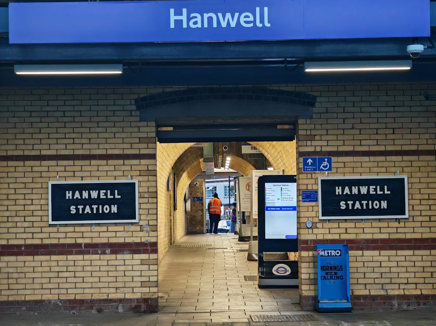 Exterior view of Hanwell Station entrance at the London Underground, featuring two black and white signs displaying 'HANWELL STATION' mounted on a beige brick wall with a red brick border. Above the entrance, a blue sign with white text indicates the station's name. The entrance has an arched brick doorway, with a glass and metal ticket machine positioned to the right, displaying station information on its screen. Inside the station, a person in an orange high-visibility jacket is visible walking away, towards the platform area. A wheelchair accessibility sign is mounted on the wall near the entrance. The scene is illuminated by overhead lighting, highlighting the brickwork and station signage, with some moving boxes and equipment nearby, suggesting recent or upcoming home relocation or furniture transport activity. This view exemplifies elements related to moving logistics, including station access and its environment.