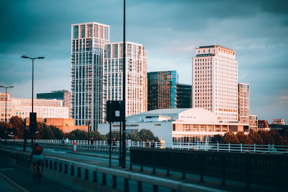 A modern urban scene featuring a London Underground metro sign with an orange background and black 'M' logo, positioned on a tall black pole near the sidewalk. Behind the sign are tall contemporary office buildings with glass facades, reflecting the sky and surrounding structures. One building displays a sign reading 'MAN SIGNATURE' at the top. The street scene includes a 30 mph speed limit sign, black metal railings along the pavement, and a few pedestrians walking past. The area appears to be a busy city centre location with a mix of commercial and transport infrastructure. This setting is relevant to house removals and moving services, illustrating access points and logistical considerations for furniture transport and home relocation near busy urban buildings, with the presence of loading zones and pedestrian pathways supporting efficient moving operations. The overall atmosphere is professional and typical of city centre environments in the UK, supporting the importance of planning for removals near building access rules and busy public spaces.