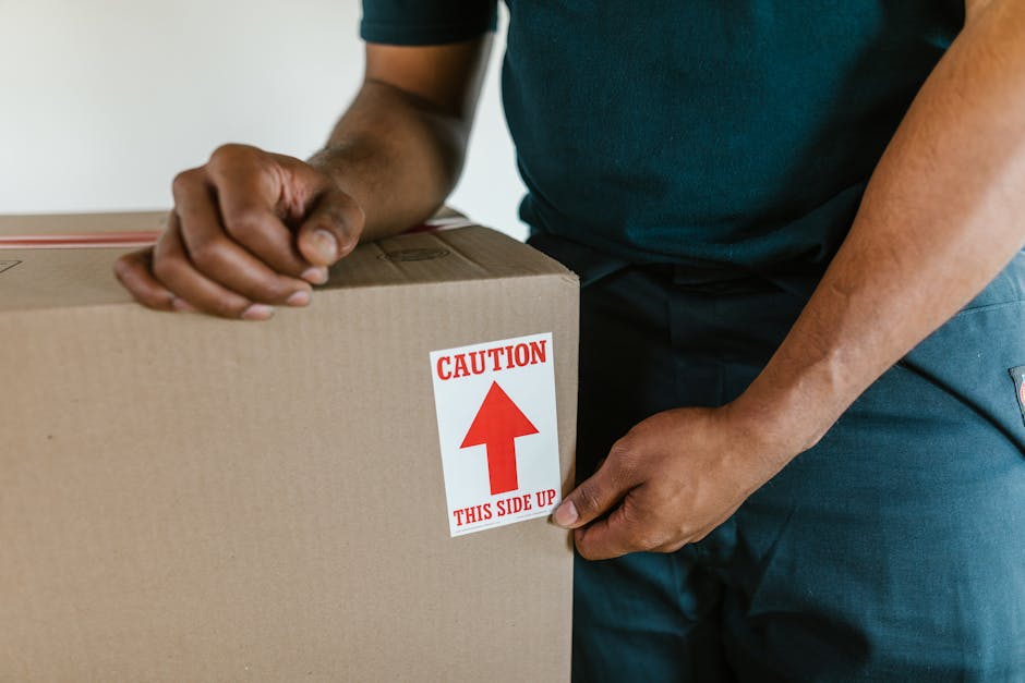 Close-up of a person wearing a dark teal shirt and dark trousers, securing a large cardboard box sealed with packing tape, with a red and white caution label that reads 'CAUTION THIS SIDE UP' and an upward arrow. The individual is holding the box steady with both hands, one on each side, preparing for transport during a home relocation process. The background shows an indoor space with neutral-colored walls, suggesting an interior environment ready for moving activities. Visible packing materials and proper handling indicate careful furniture transport and packing practices typical of professional removals services such as those offered by removalvanelephantandcastle.co.uk. This image captures the loading process, emphasizing safe handling and proper packing techniques crucial for smooth furniture transport during house removals on New Kent Road, Elephant and Castle.