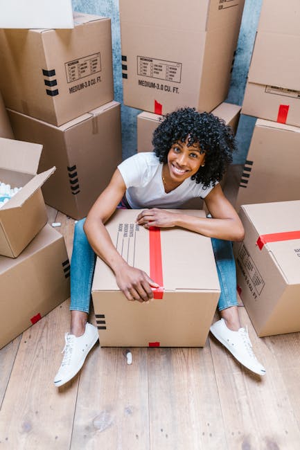 A woman with curly black hair and a white t-shirt is sitting on a wooden floor surrounded by numerous cardboard boxes, some sealed with red packing tape and others open with packing materials visible inside. She is smiling at the camera while gently holding a large sealed box in front of her. The boxes vary in size, with some stacked vertically against a blue wall, indicating an indoor space prepared for home relocation. The environment appears well-lit, likely in a residential setting, and the scene captures a moment during packing or loading in the house moving process, reflecting careful handling of household items, which is typical for professional removals like those offered by [COMPANY_NAME]. The overall scene emphasizes packing, organisation, and preparation for a furniture transport or home move, aligning with the themes of the page about top tips for Elephant and Castle moves.