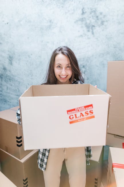 Close-up of a person wearing a dark teal shirt and dark trousers, securing a large cardboard box sealed with packing tape, with a red and white caution label that reads 'CAUTION THIS SIDE UP' and an upward arrow. The individual is holding the box steady with both hands, one on each side, preparing for transport during a home relocation process. The background shows an indoor space with neutral-colored walls, suggesting an interior environment ready for moving activities. Visible packing materials and proper handling indicate careful furniture transport and packing practices typical of professional removals services such as those offered by removalvanelephantandcastle.co.uk. This image captures the loading process, emphasizing safe handling and proper packing techniques crucial for smooth furniture transport during house removals on New Kent Road, Elephant and Castle.