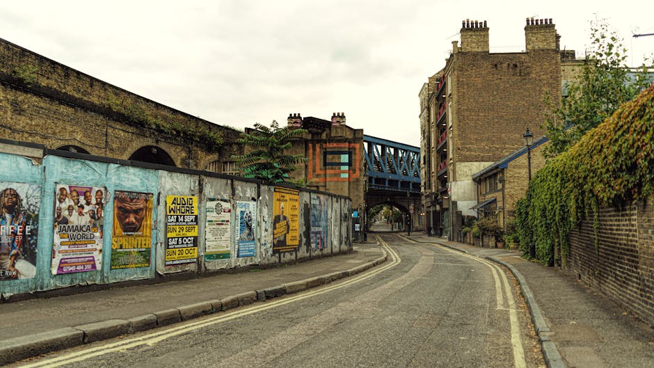 A narrow urban street scene with a curved asphalt road leading towards a bridge with blue metal framework and brick buildings, some with multiple chimneys, on both sides. On the left, a long weathered wooden fence covered with colorful posters advertises various events, including music and community gatherings, while the right side features brick walls partially covered with green ivy and foliage. The street is empty of vehicles and pedestrians, and the overcast sky creates diffuse lighting. The scene depicts an area suitable for home relocation or furniture transport services, with visible pavement and a typical London-style environment, aligning with the relocation services offered by removalvanelephantandcastle.co.uk.