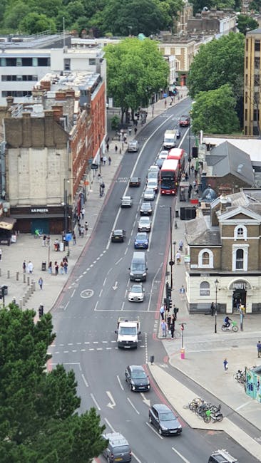 An aerial view of Walworth Road in Elephant and Castle showing a busy street with multiple vehicles including cars, a red double-decker bus, and vans, moving along the road. The scene includes a mixture of commercial and residential buildings, with some featuring brick facades and others modern construction. Trees line the sidewalks, providing greenery amidst the urban environment. Pedestrians are walking on the pavements, and bicycles are parked near the corner. The image captures the typical setting for home relocation and furniture transport logistics, highlighting the street's activity during the day which may be relevant for planning removals or moving services offered by [COMPANY_NAME] as part of a comprehensive house moving process.